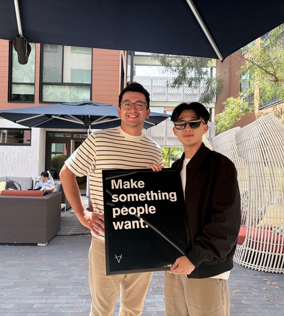 Two teammates in a courtyard holding a framed “Make something people want” poster.